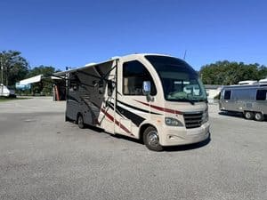 Modern RV with awning extended in an open lot under clear blue sky.