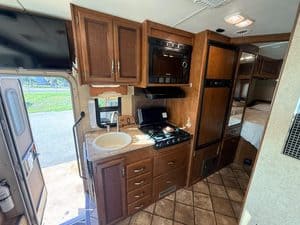 RV kitchen with wooden cabinets, microwave, stove, and sleeping area in background.