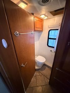 Small RV bathroom with toilet, window, and wooden cabinets.