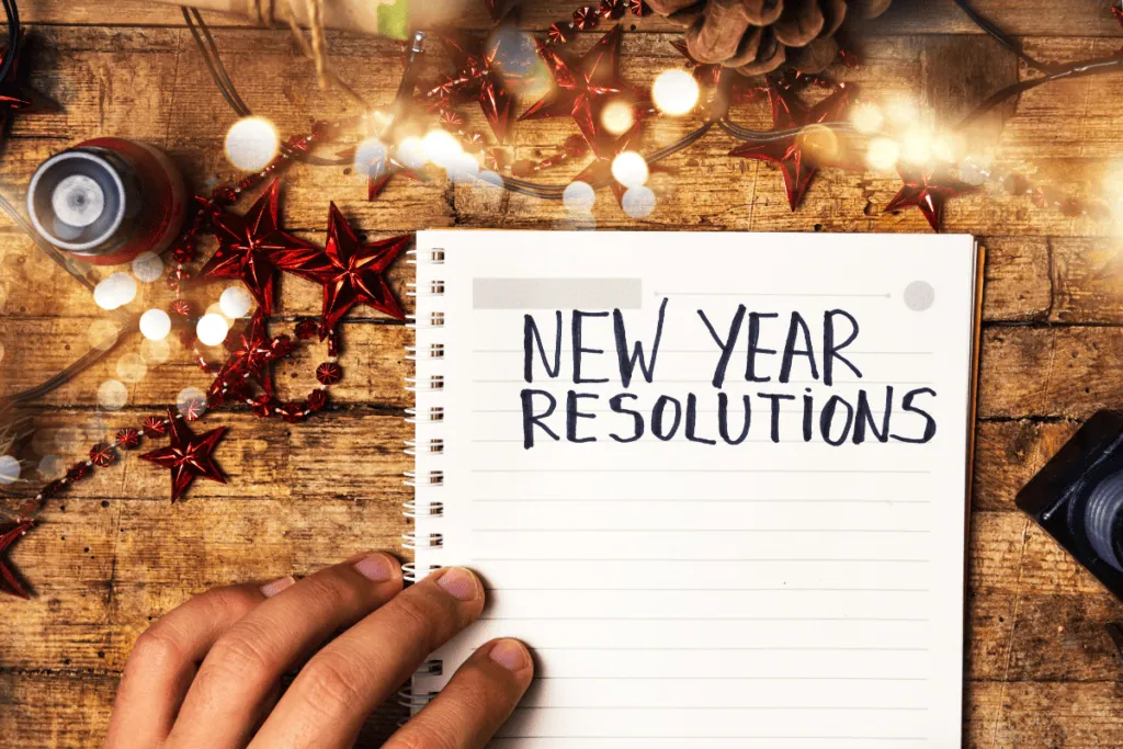 An open notebook on a wooden table with the words “New Year Resolutions” written on the page.