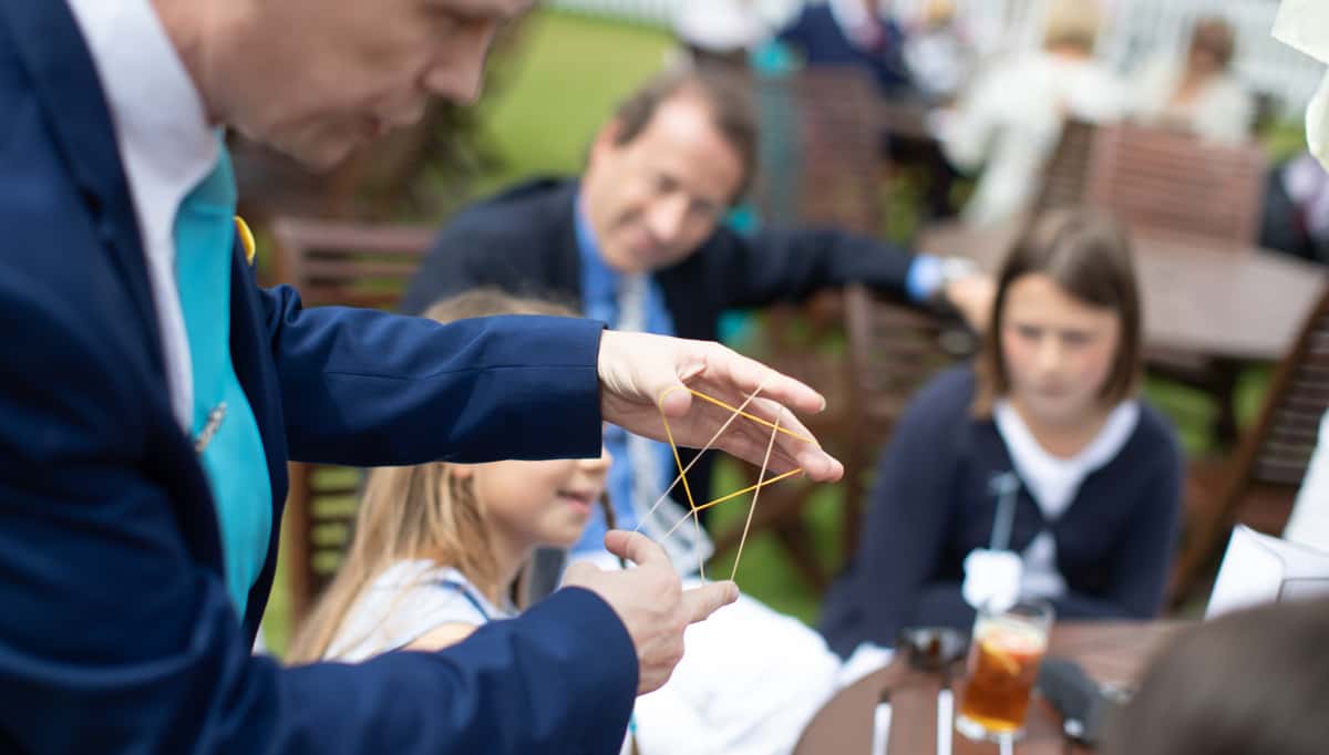 A person performs a rubber band magic trick for a group of children and adults seated at an outdoor table.