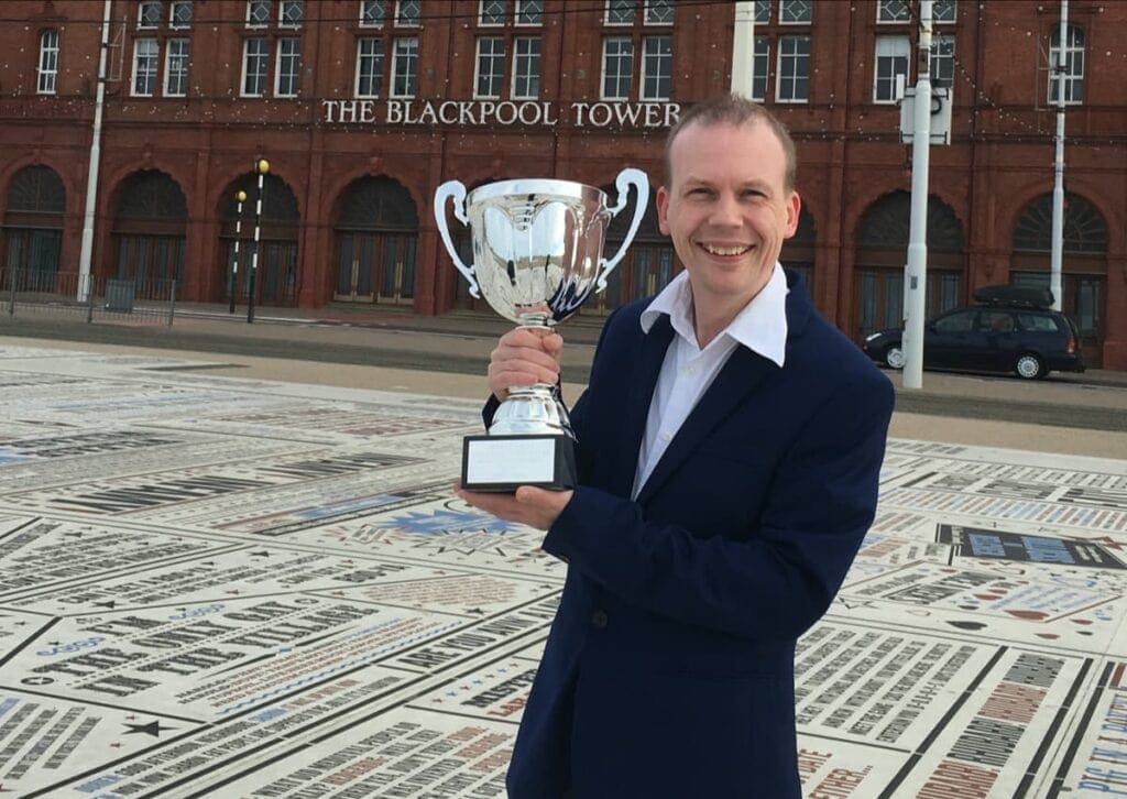 A person in a suit stands outside The Blackpool Tower holding a large silver trophy and smiling at the camera.