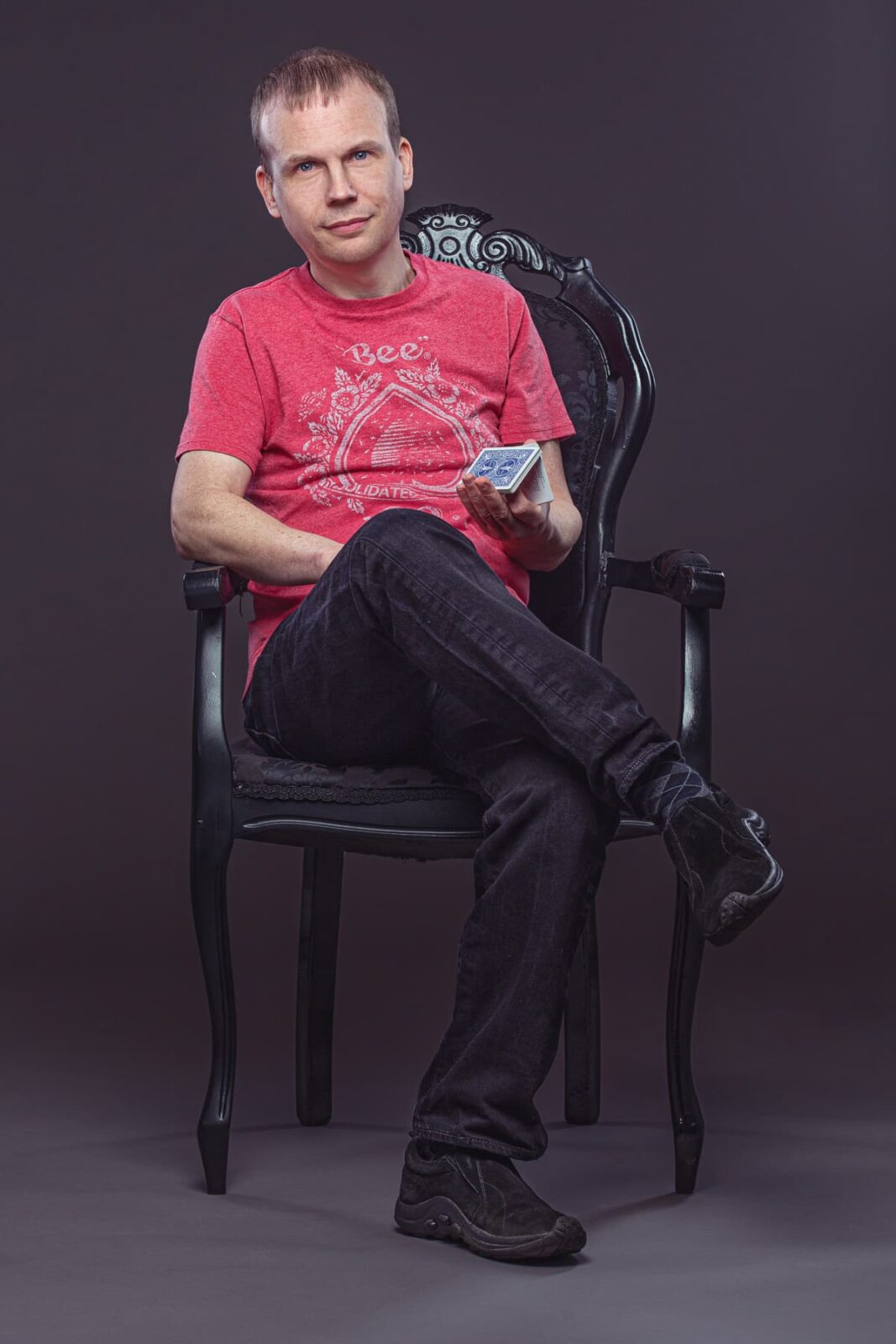 A man in a red graphic T-shirt and black jeans sits on a black ornate chair, legs crossed, holding a pack of cards in one hand against a plain dark background.