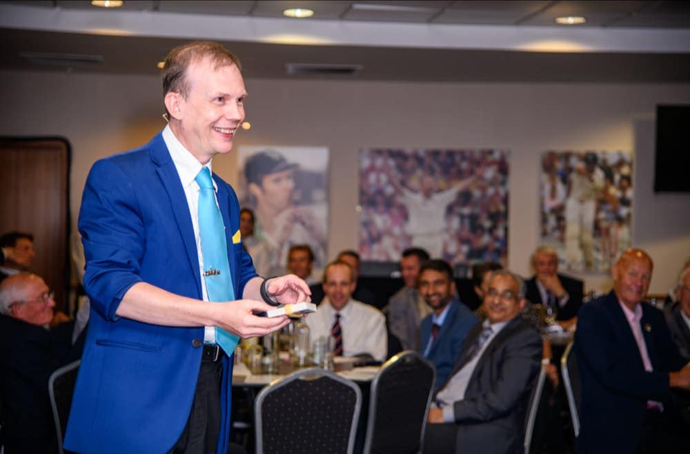 A man in a blue suit jacket performs a card trick in front of an audience seated at tables in a well-lit function room.
