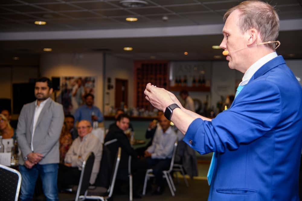 A man in a blue suit speaks into a headset microphone whilst addressing an audience seated at tables in a conference room.