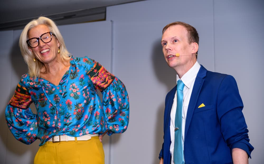 Two presenters stand on stage; the woman wears a floral blouse and yellow trousers, smiling, while the man in a blue suit and tie looks ahead. Both have headset microphones.