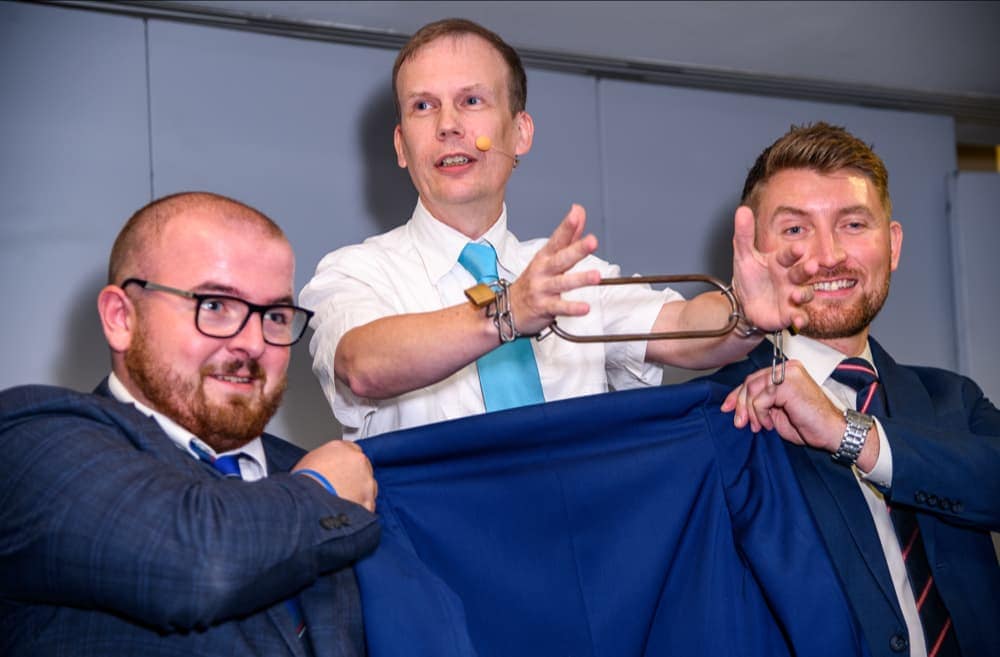 Three men in suits, one performing a handcuff trick with metal restraints, while the other two hold a blue jacket in front of him.