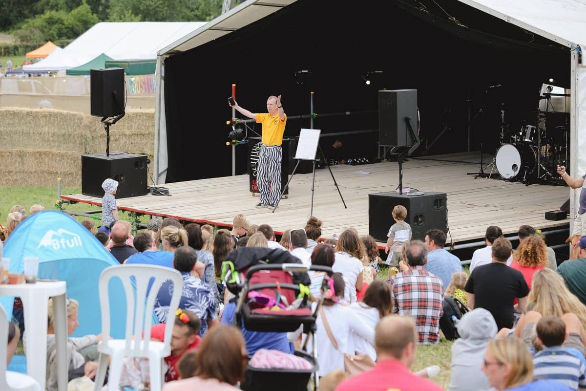 A performer in a yellow shirt and striped trousers entertains a seated audience on an outdoor stage at a festival or event.