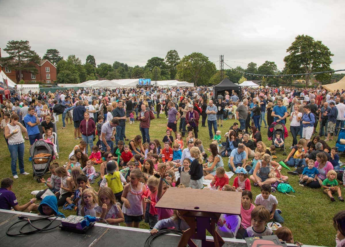 Large crowd gathered at an outdoor festival with families and children seated on the grass and people standing near white marquees on a cloudy day.