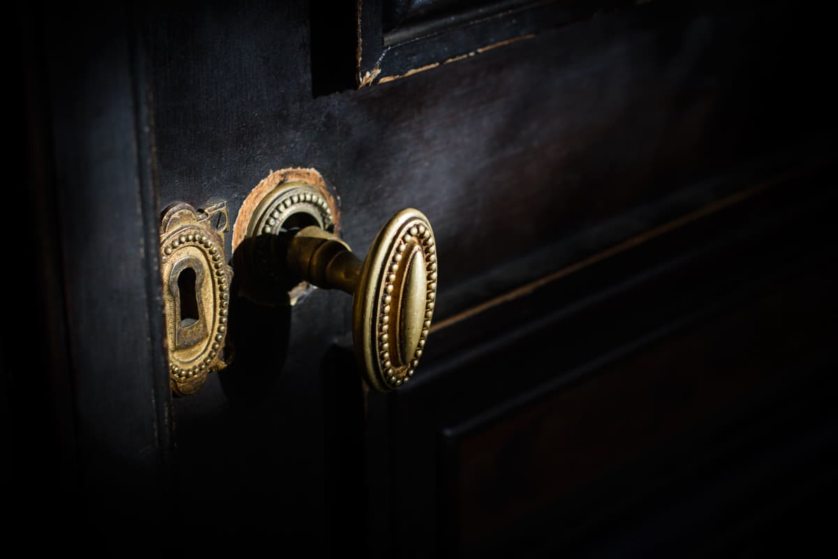 Close-up of a vintage brass door handle and keyhole on a dark wooden door, illuminated by focussed light, with signs of wear around the keyhole.