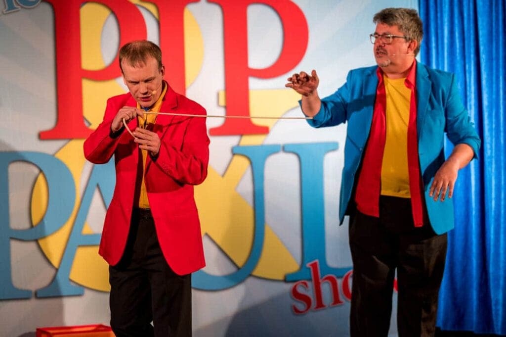Two men in colourful jackets perform a magic trick with a long white string on stage in front of a backdrop that reads "Pip and Pakki Show.