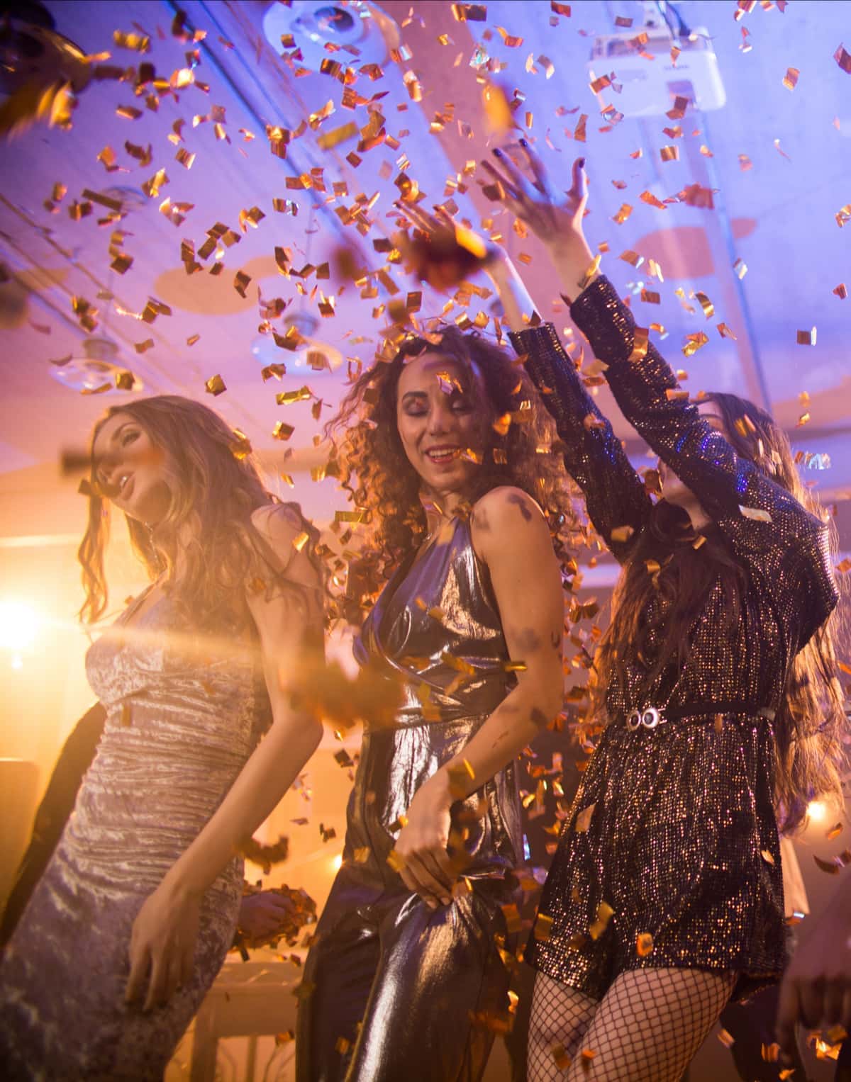Three women in shiny dresses dance and celebrate as gold confetti falls around them at a party, with warm lighting in the background.