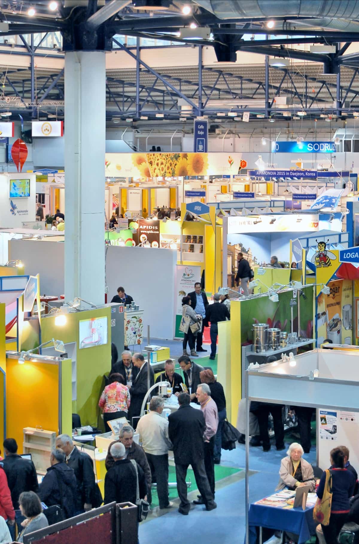 Crowds of people walk and interact at an indoor trade show or exhibition, with multiple stands and colourful displays visible throughout the large hall.
