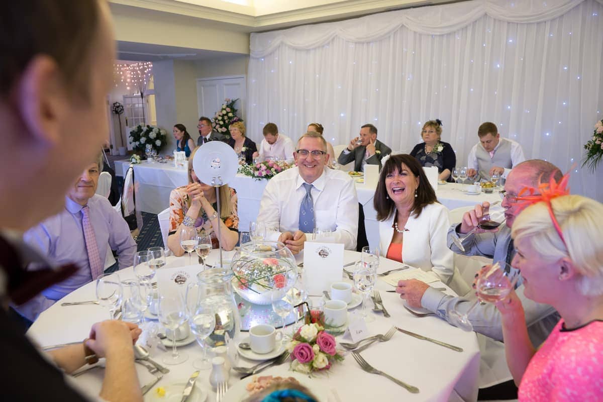 A group of people sit around a decorated table at a formal event, smiling and laughing, with one person covering their face with a menu.