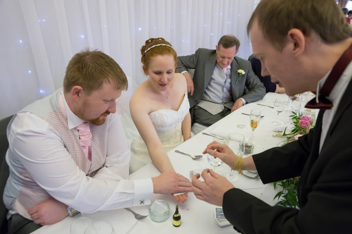 A bride and groom watch as a man in a suit performs a magic trick at a wedding reception; another guest sits in the background.