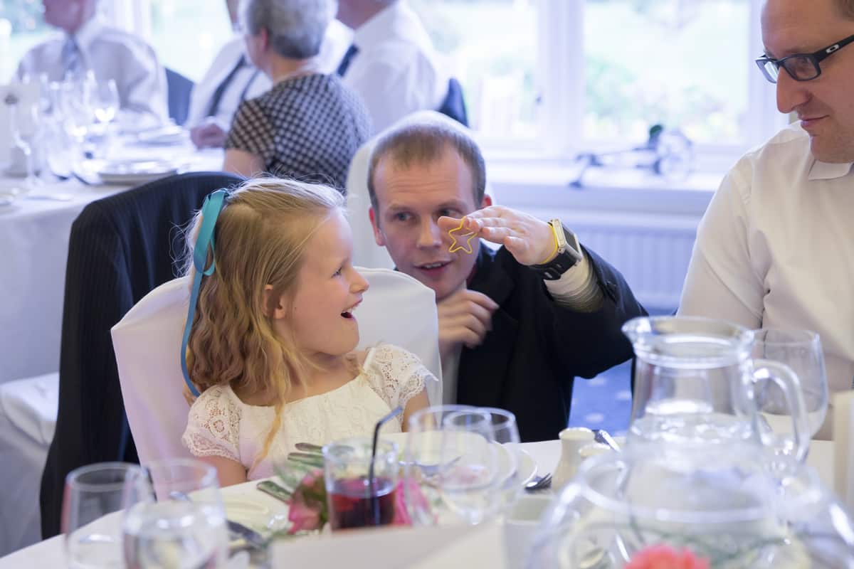A man shows a star-shaped object to a smiling girl at a formal table laid for a meal, with other people seated in the background.