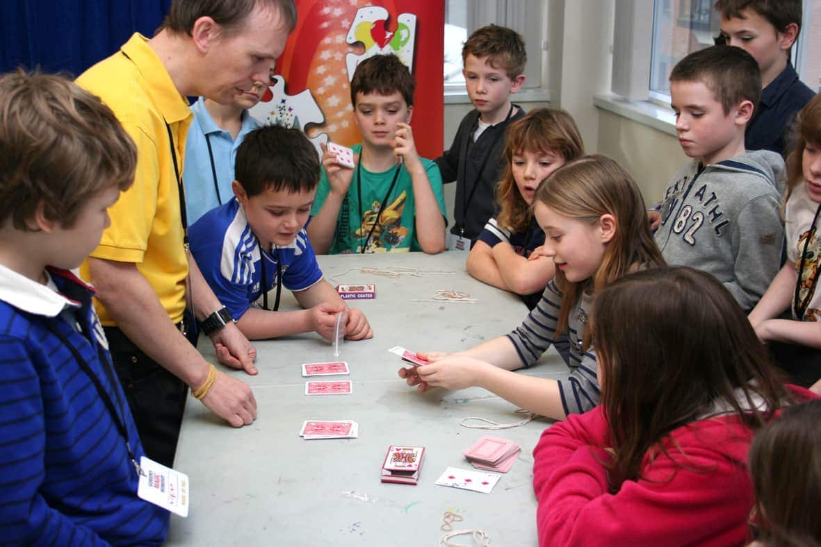 A group of children sit and stand round a table as a man in a yellow shirt leads a card game, with playing cards and elastic bands on the table.