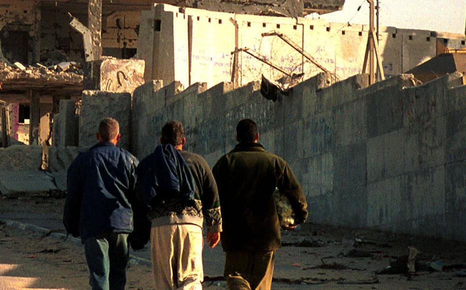 Palestinians walk to Khan Younis December 10, 2001 after crossing a checkpoint into an Israeli settled area in the Gaza strip. A crane on the Israeli side builds up a wall around the settlement. In the background are buildings in Khan Younis that Israeli forces have fired into.