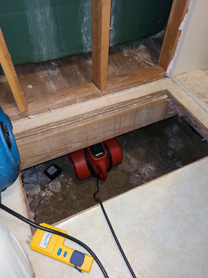 Red air mover set into an opened under-floor cavity to dry the crawlspace, with a moisture meter in the foreground, during water damage mitigation in Denison, TX