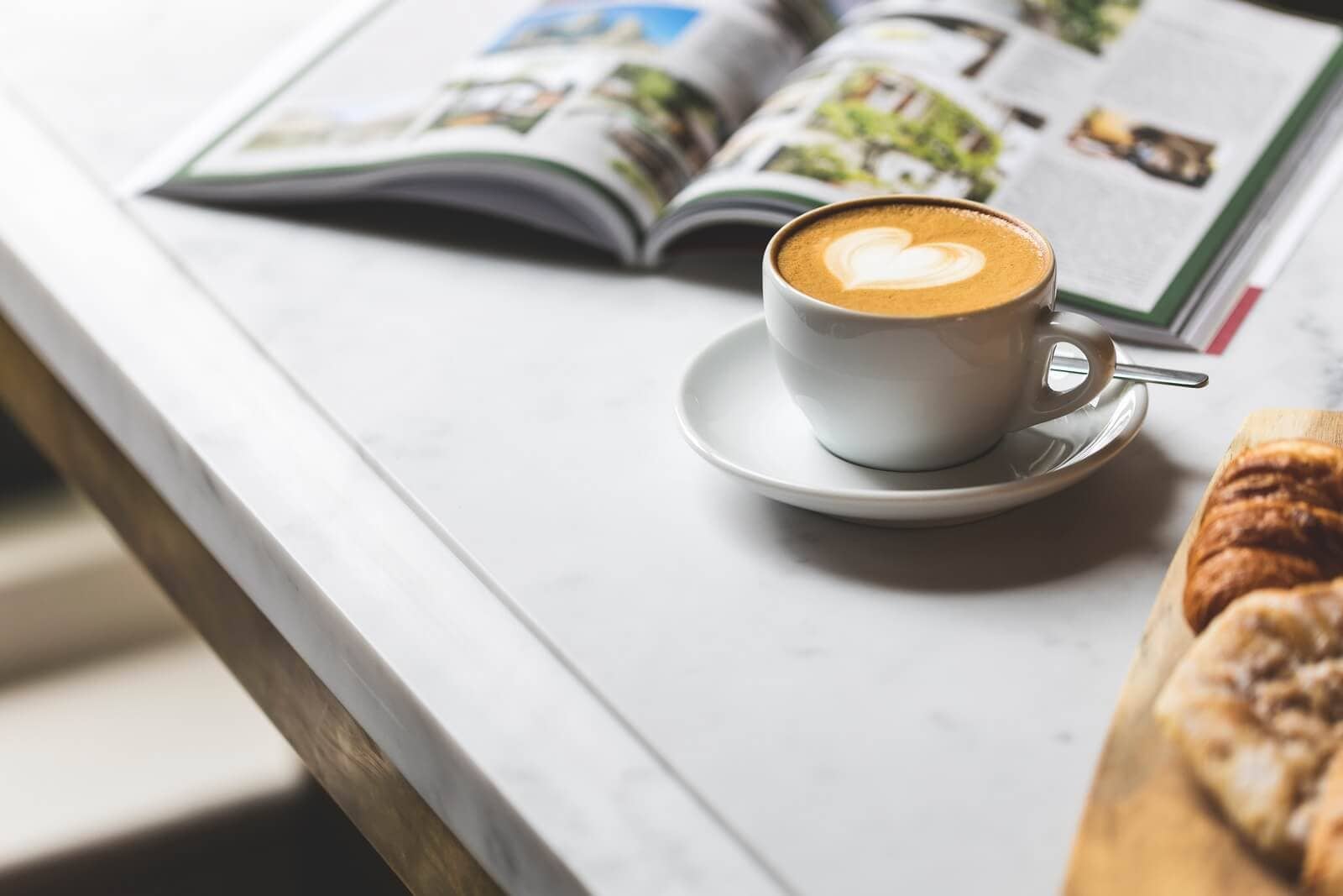 Kaffeetasse mit Herz-Milchschaum, Frühstück und Magazin auf Tisch.