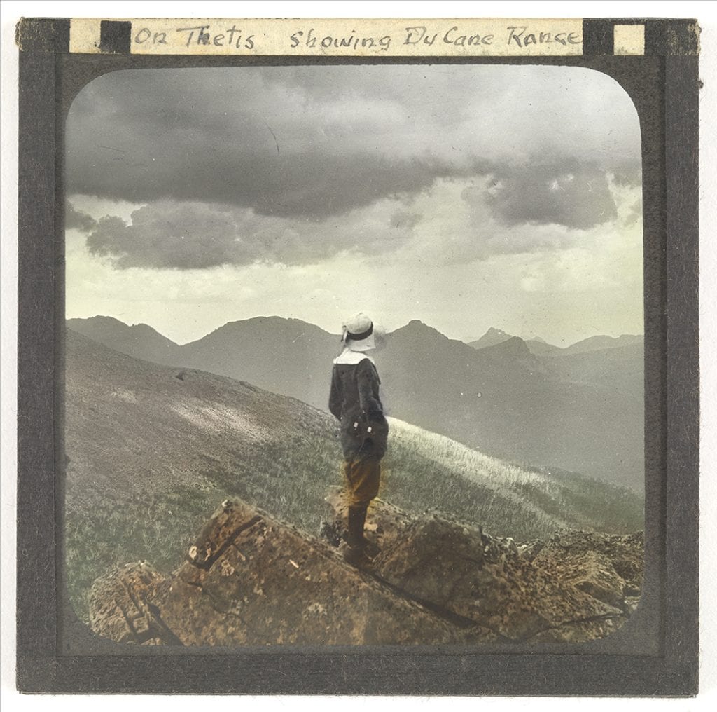 Photograph - On Thetis showing Du Cane Ranbfe [Female bushwalker standing on clifftop, watching sky and mountain tops in distance] [Lantern Slide] [Box 2]