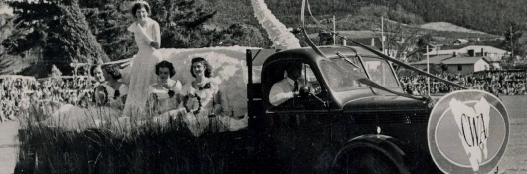 Black and white image of a parade float with several young women dressed in white next to a sculpture of a swan. A man drives the truck. A sign bearing the CWA Tasmania logo attached to the front of the truck. A crowd and fairground are in the background.