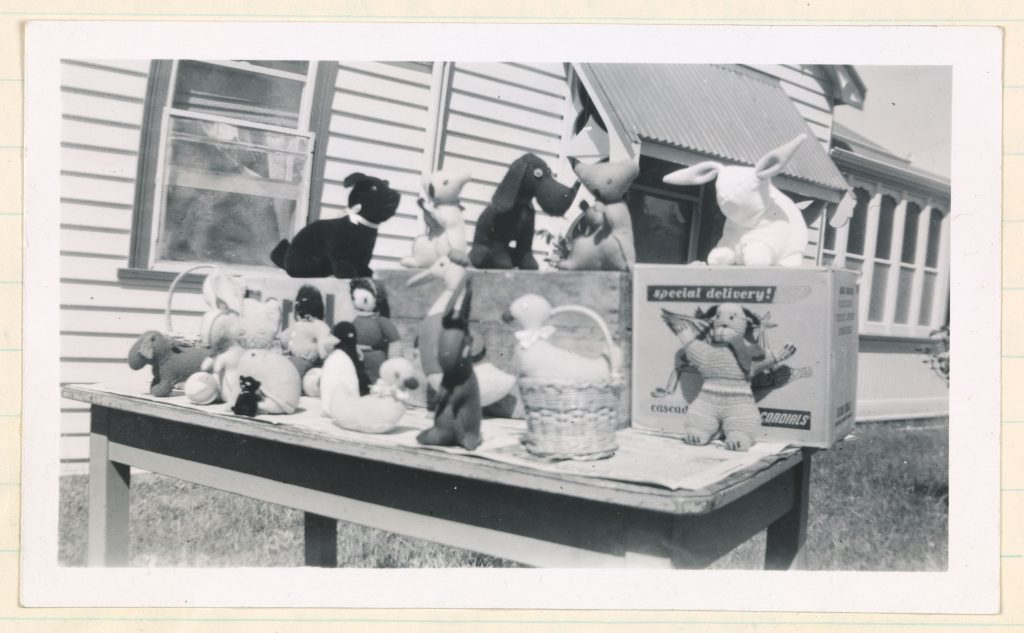 Black and white photograph of a table displaying handmade stuffed toys outside a weatherboard building.