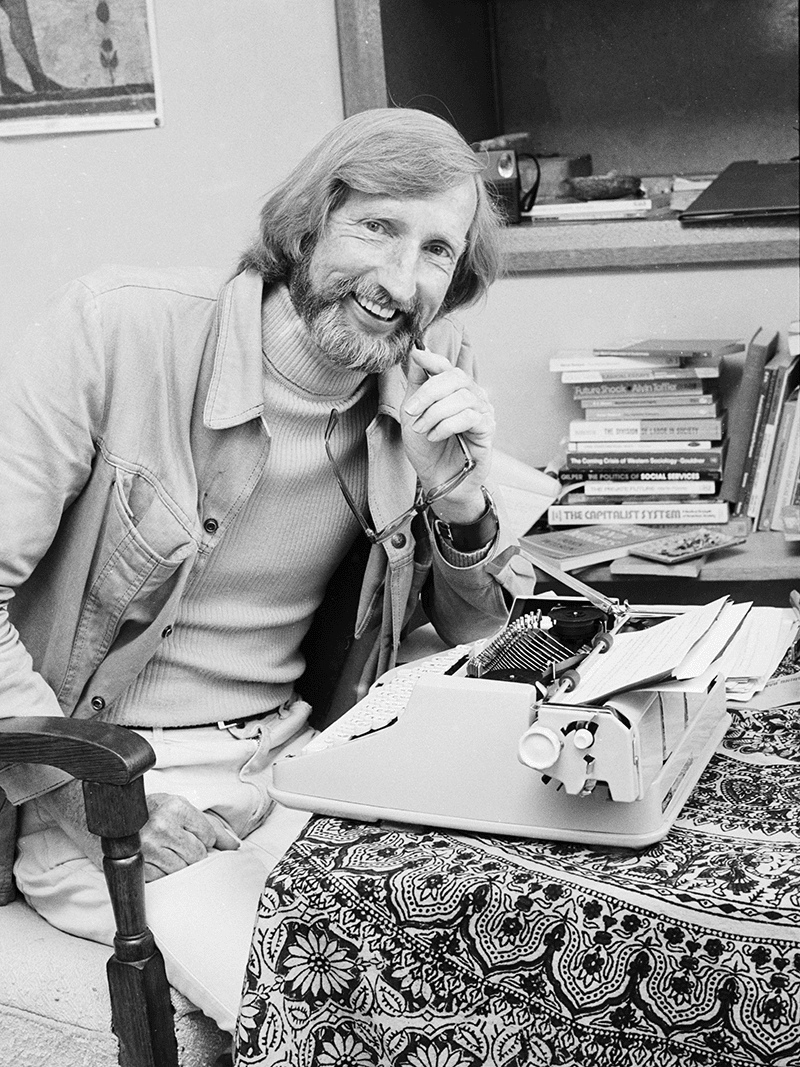 Image of Roy Bridges, an elderly man smiling while sitting in front of a typewriter at a desk with a patterned tablecloth. A stack of books sits on a table behind him.