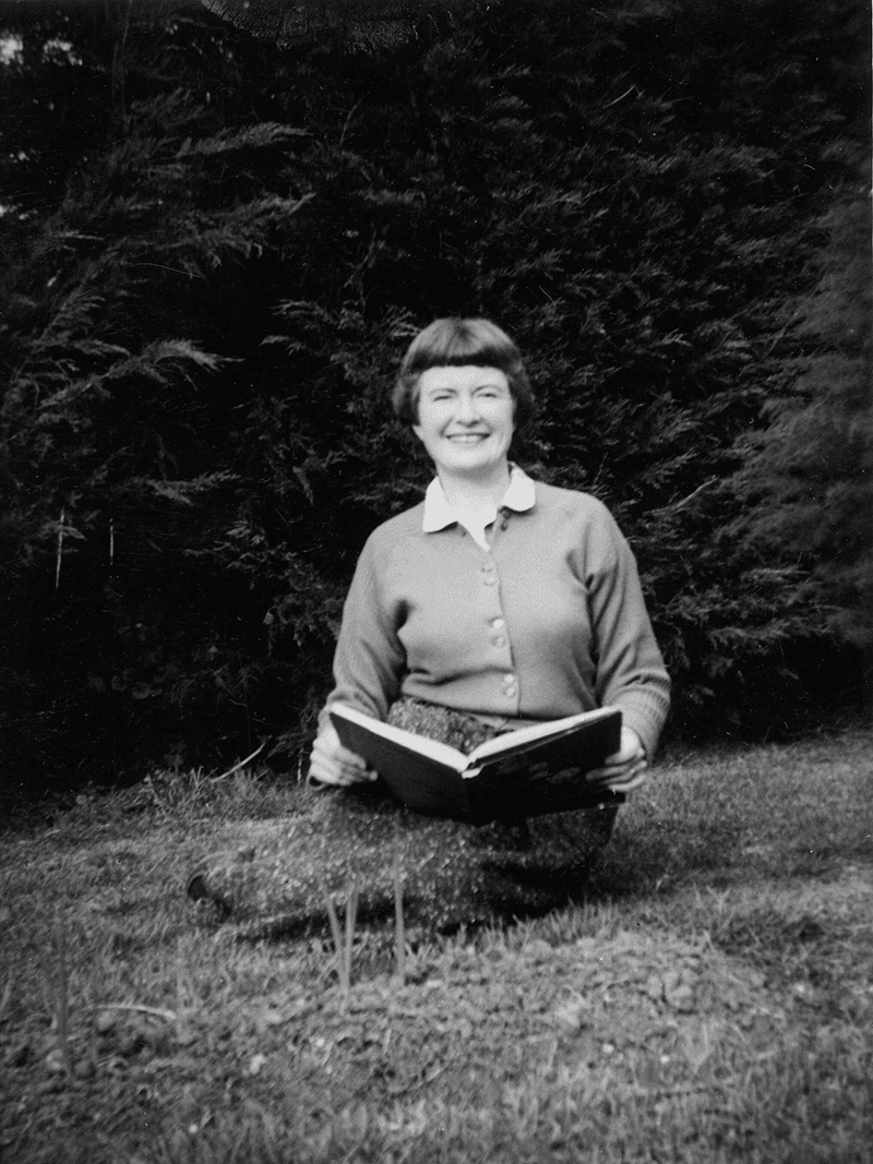 Image of Gwen Harwood smiling while sitting on the grass with an open book in her hands.