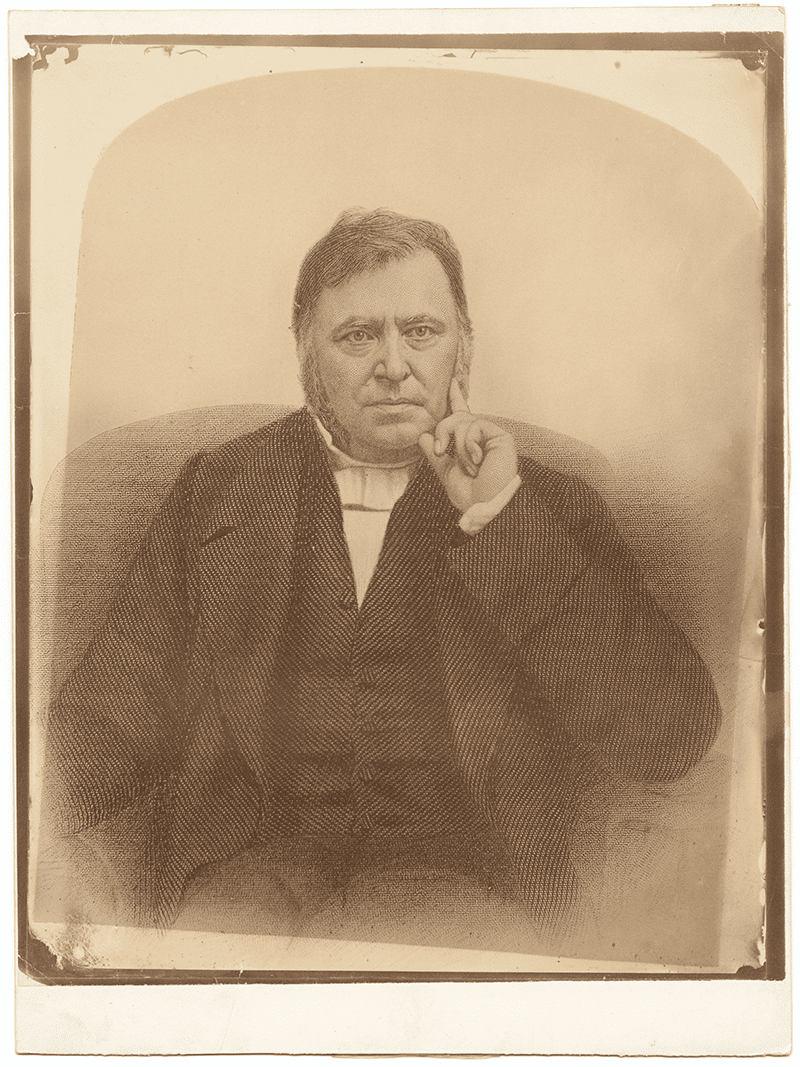 Portrait of John West, a middle-aged man with sideburns, wearing a dark suit and white cravat. He sits with his right hand near his face, index finger resting on his cheek