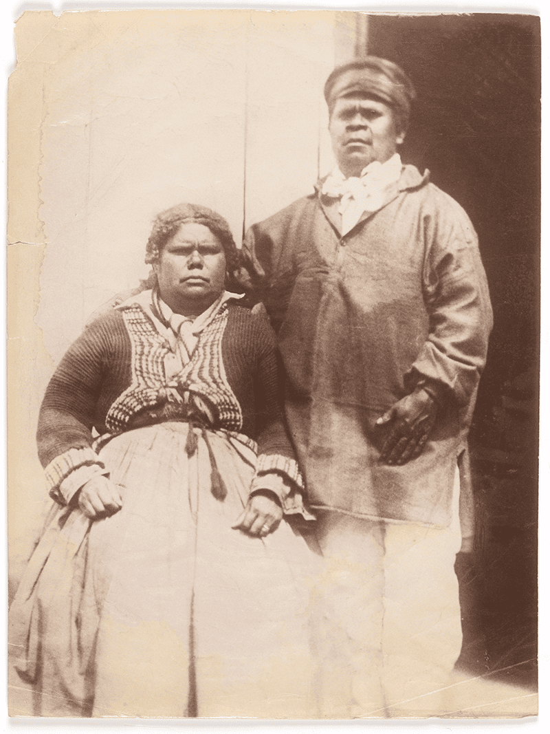 Image of Mary Ann Arthur (Clarke) and Walter George Arthur at Putalina/Oyster Cove. Two Indigenous people, a seated woman in a patterned blouse and long skirt, and a standing man in a loose shirt and hat.