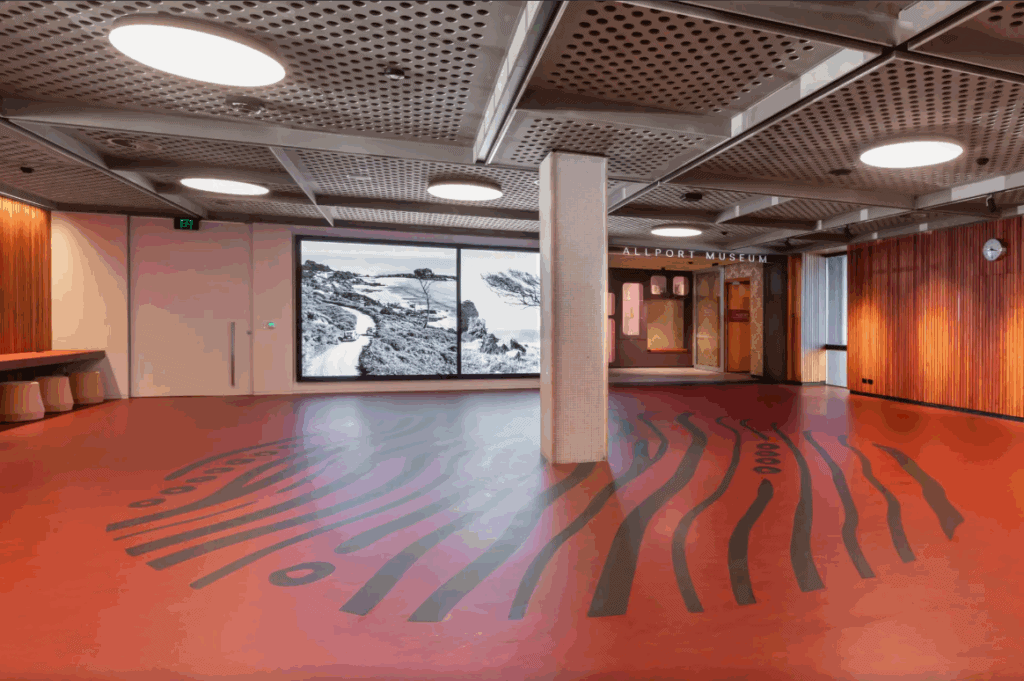 The Hobart Library ground floor. A black circular floor pattern made of lines surrounds a pillar with a big presentation screen in the background. The ceiling is grey metal with grated holes. The rest of the floor is orange lino.