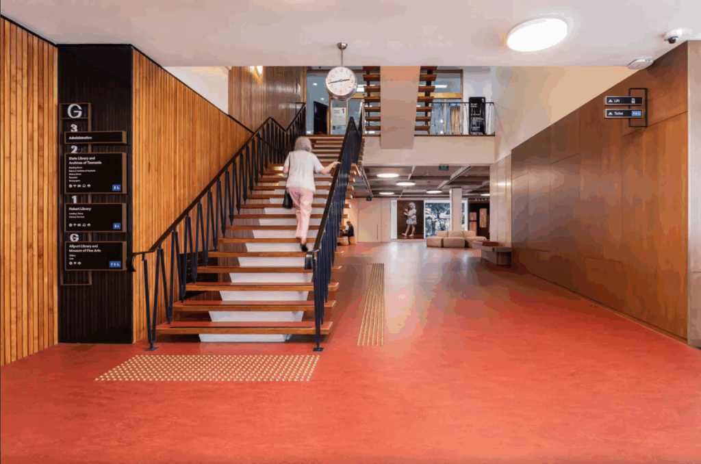 The Hobart Library ground floor. A wide hallway with a staircase on the left. Wood panel walls and an organ pattern lino floor. A clock hangs from the white ceiling.