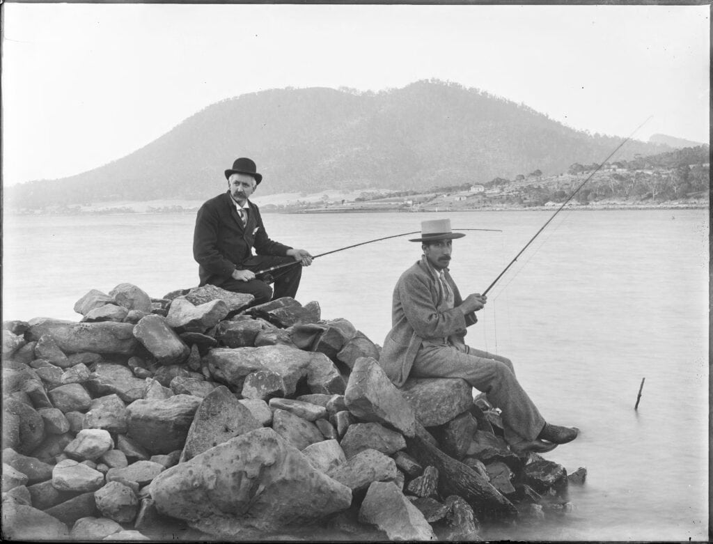 Two men in suits sitting on rocks next to a river fishing.