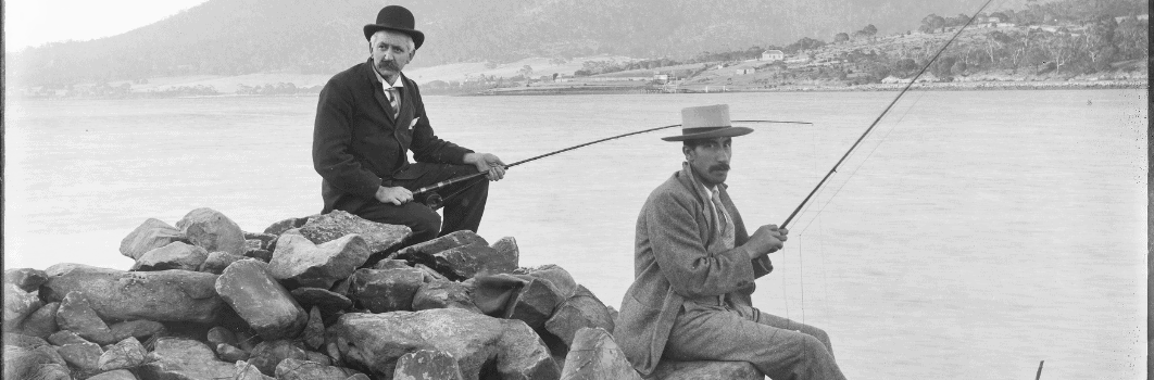 A black and white image of two men sitting on a rocky shore holding fishing rods