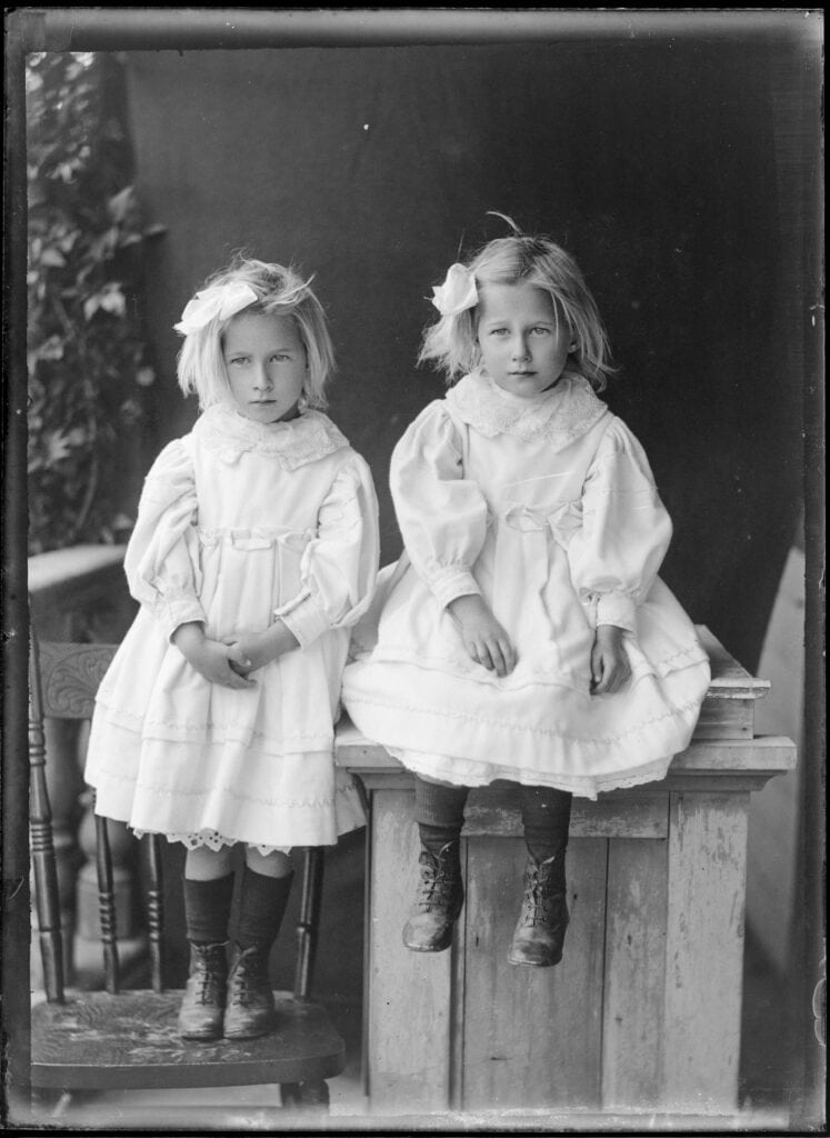 Two young twin girls in white dresses posing for a phot. One stands on a chair, the other sits on a table.