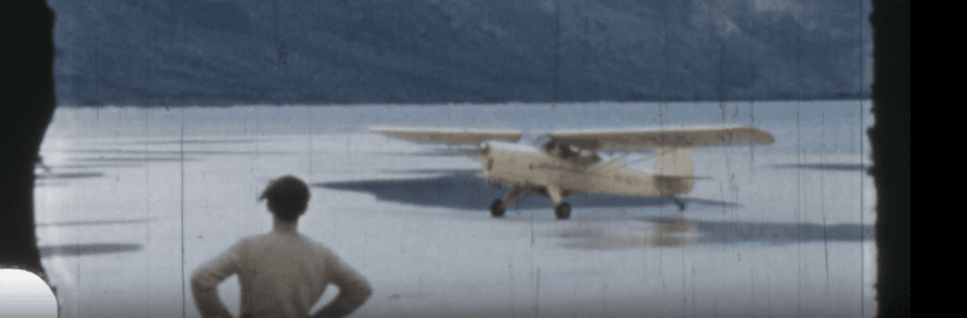 Man with hands on hips looks at yellow Auster plane that has landed on the sandy alpine beach of Lake Pedder