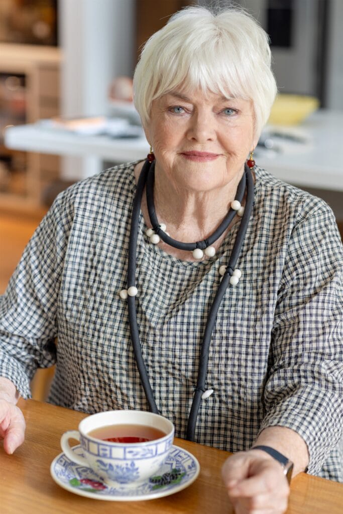 Elderly smiling woman with white hair wearing a navy checkered shirt, sitting at a table with a blue and white patterned teacup filled with tea.