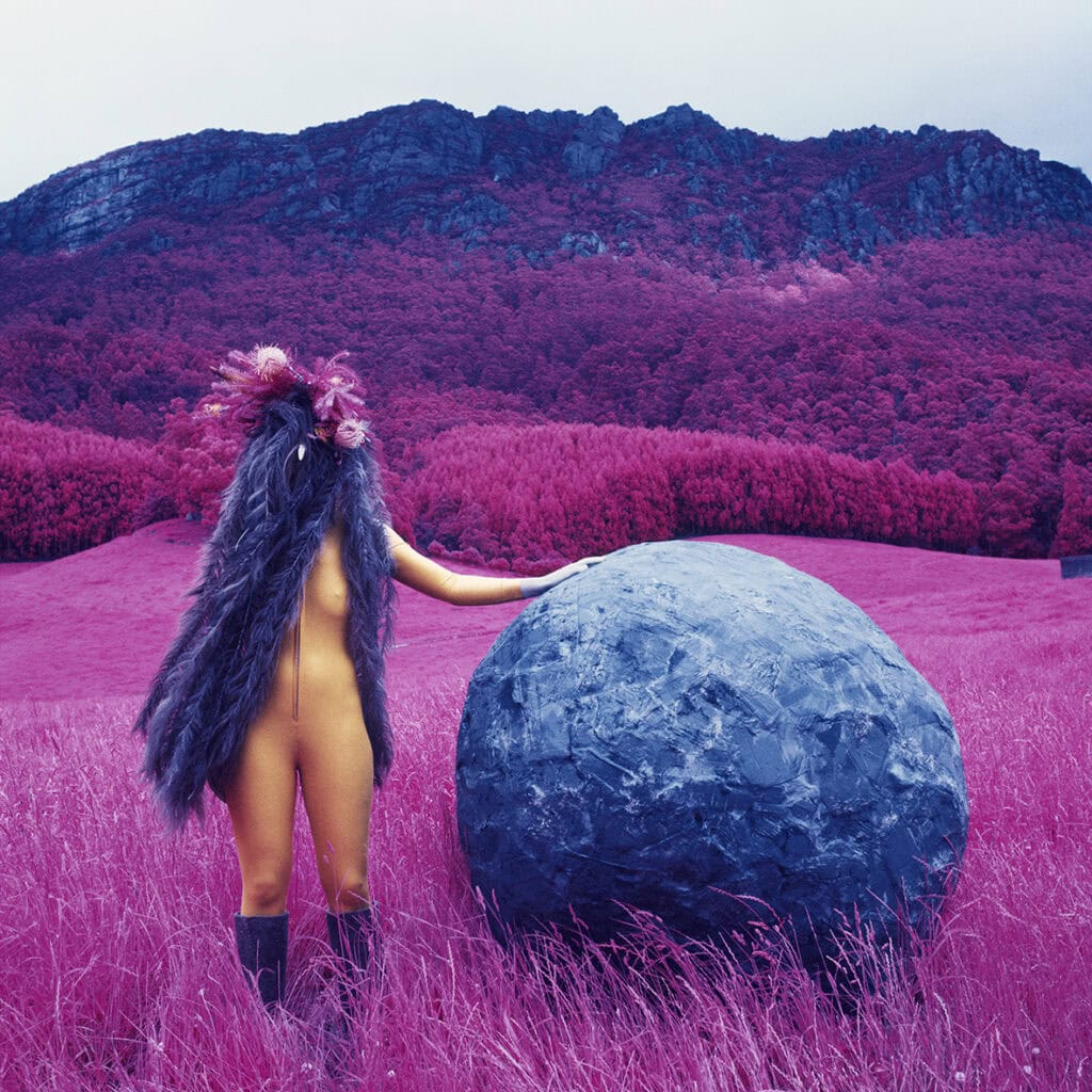A costumed woman in a skin-tight, full-body yellow suit, wearing a long purple wig with purple flowers at the top. The person stands in a field next to a boulder. The background are dense trees stretching towards a mountain. The green in the image has been colour shifted to brights pink.