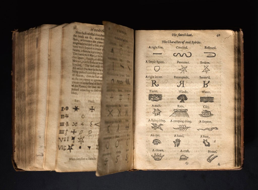 An open antique book with worn, yellowed pages sits against a dark background, its pages slightly fanned. The visible spread is titled “The fourth book. The Characters of evil Spirits,” showing rows of small labelled diagrams and symbols—lines, shapes, letters, and simple illustrations like flame, wind, water, and rain.