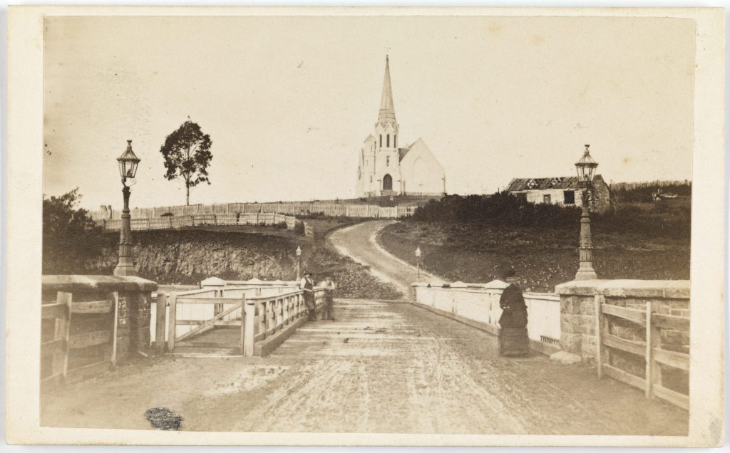 An old black and white image of a bridge with a road leading to a church in the background