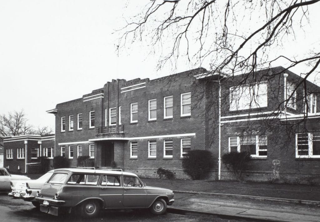 Black and white photo taken in the 1970s of the front view of an extensive two storey brick building.  Cars are parked in front of the main entrance.