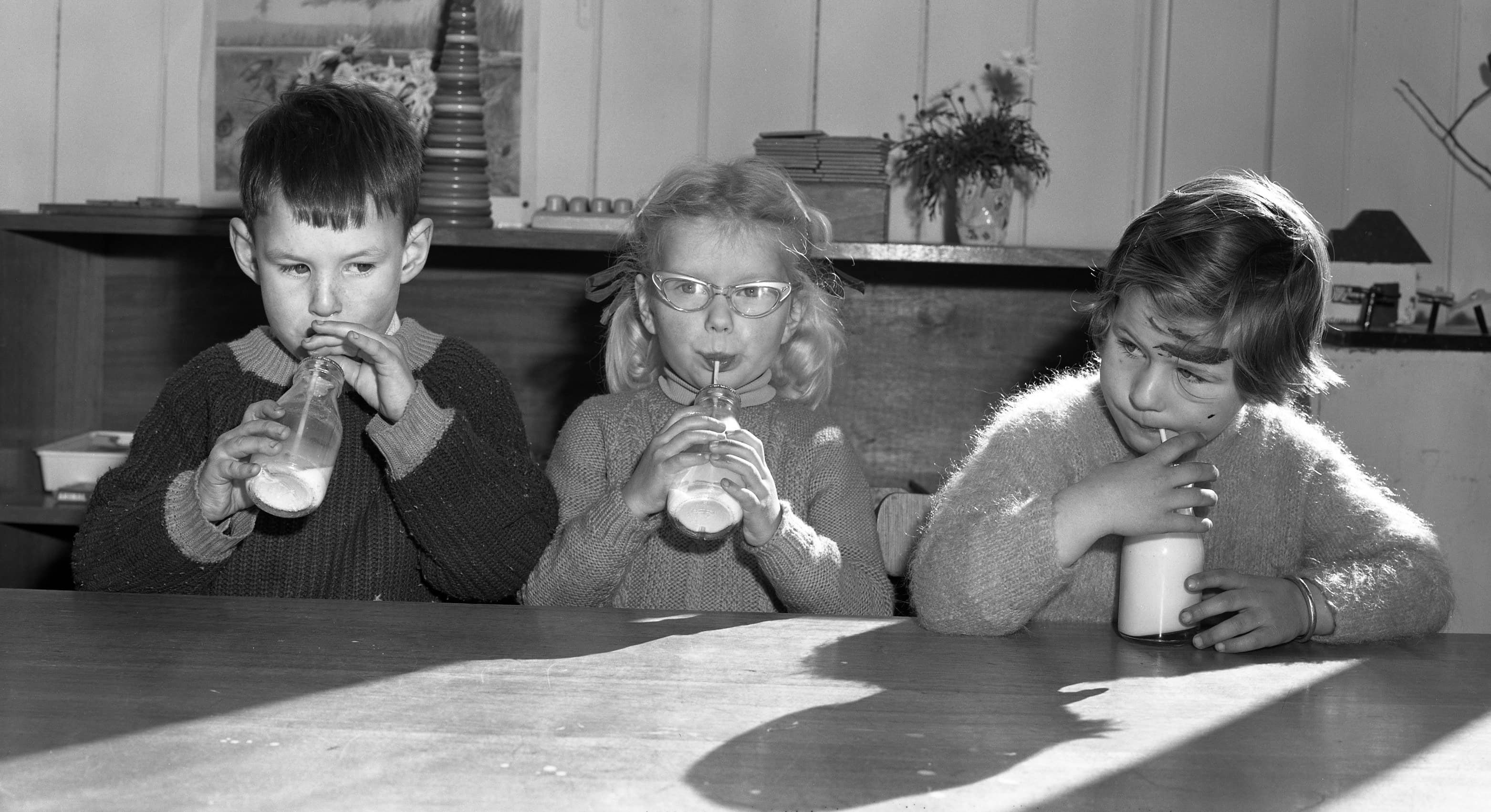 a black and white photo of 3 children drinking milk at a table