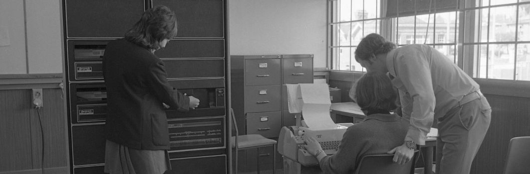 an old black and white photo of 3 students using computer with a ticker tape printout