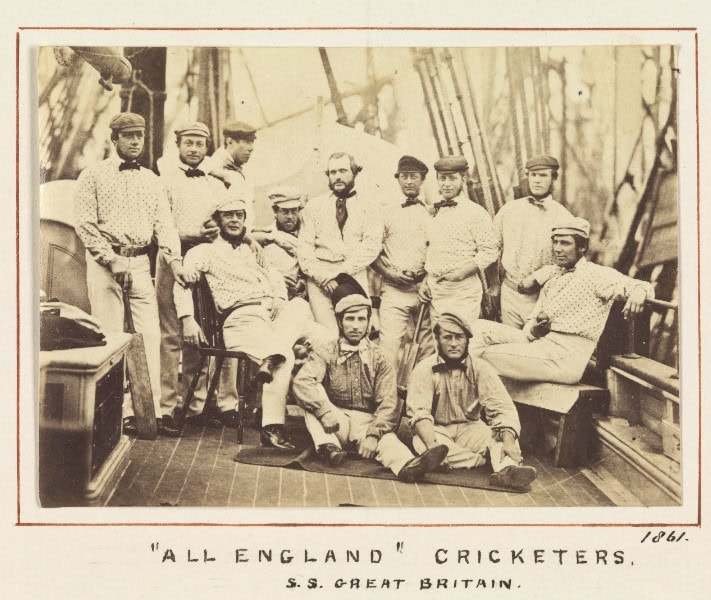 A black and white photo of 12 men in cricketer uniforms posing for a photo on the deck of a sailing ship.