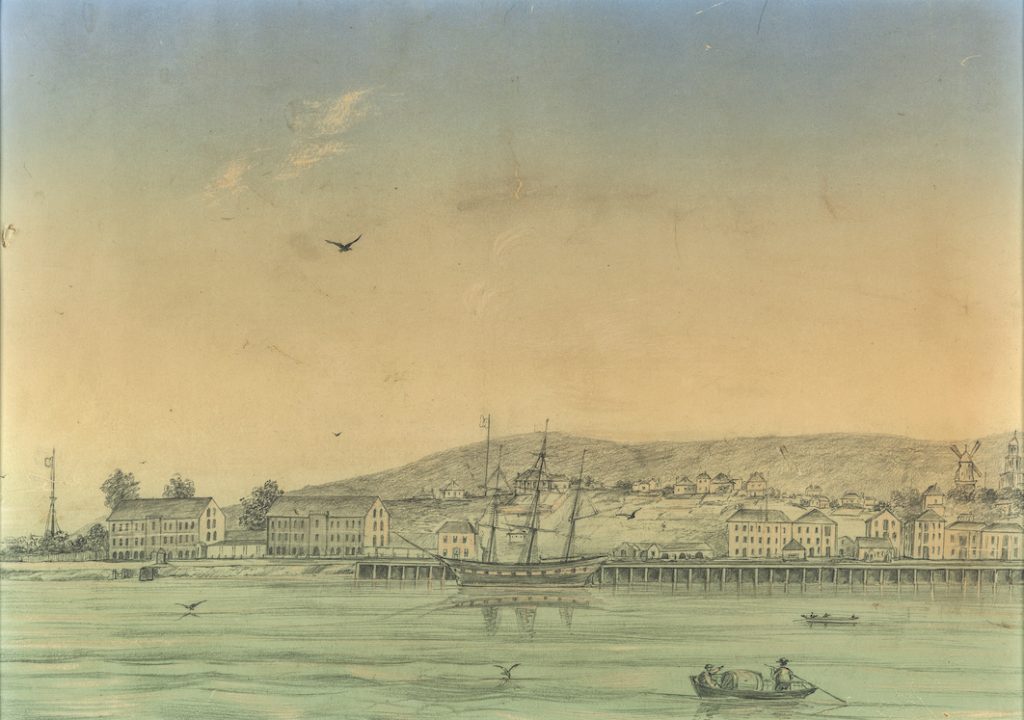 A painting of a town viewed from the water. a ship and boat can be seen in the foreground. Mountains in the distance.