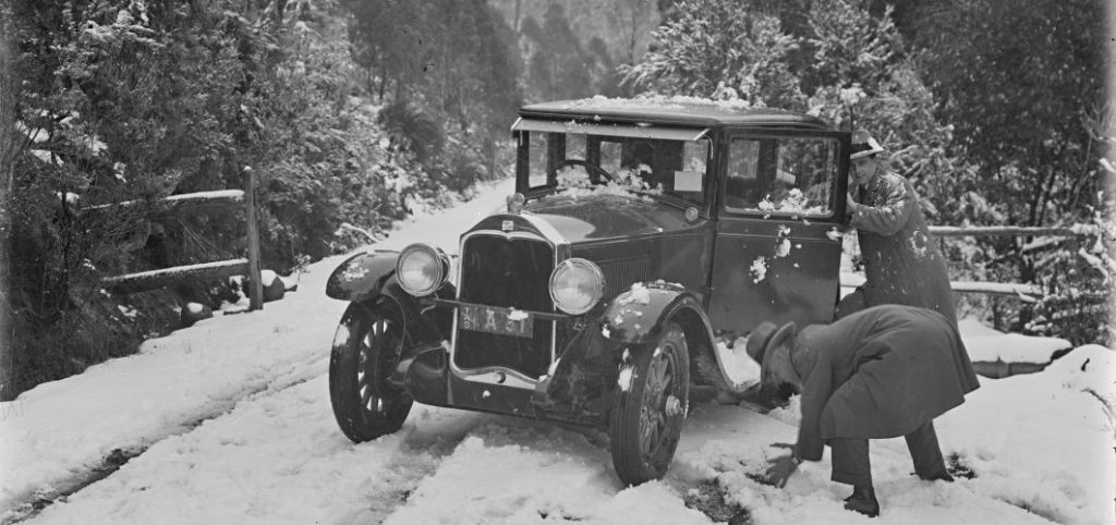 A black and white photo of 2 men stopping to admire the snow next to an old car.