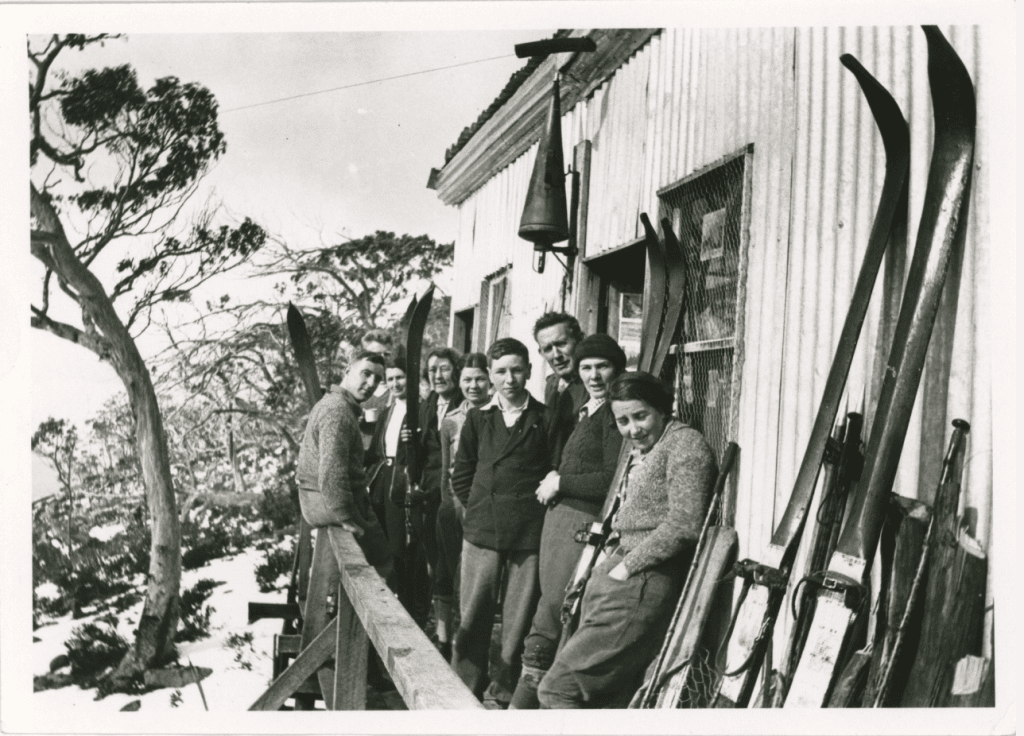 An old black and white photo of 9 people standing on the balcony of a building known as "Mulga Micks's hut." with their skis. 