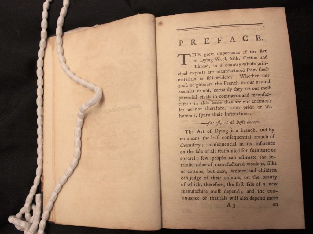 A n open book. the text reads: “Preface. The great importance of the art of dying wool, silk, cotton and thread, in a country whole principal exports are manufactured from these materials is self-evident. whether our good neighbours the French be our natural enemies or not, certainly they are our most powerful rivals in commerce and manufactures: in this sense they are our enemies; let us not therefore from pride or ill-humour, spurn their instructions. The art of dying is a branch, and by no means the least consequential branch of chemistry consequential in its influence on the sale of all stuffs used for furniture or apparel: few people can estimate the intrinsic value of manufactured woollens, silks or cottons, but men, women and children can judge of their colours, on the beauty of which therefore, the fist sale of a new manufacture must depend; and the continuance of that sale will also depend more on.”