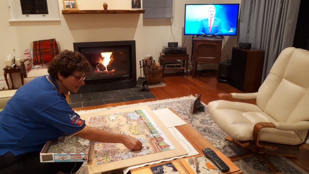 A picture of a person doing a jigsaw puzzle in a living room with the tv on in the background.